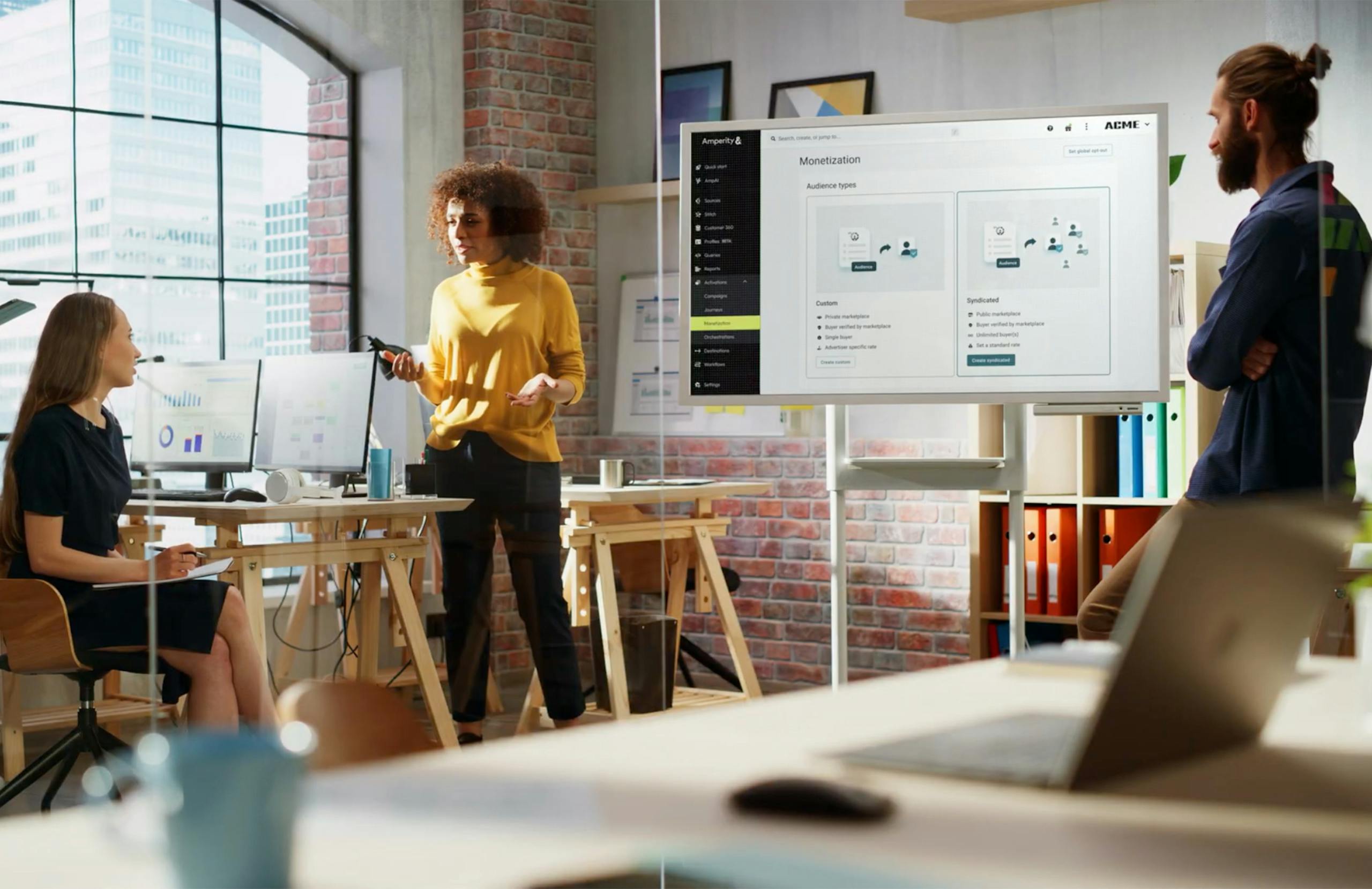 Three co-workers collaborating in a brick office with a screen of Amperity Monetization types displayed.