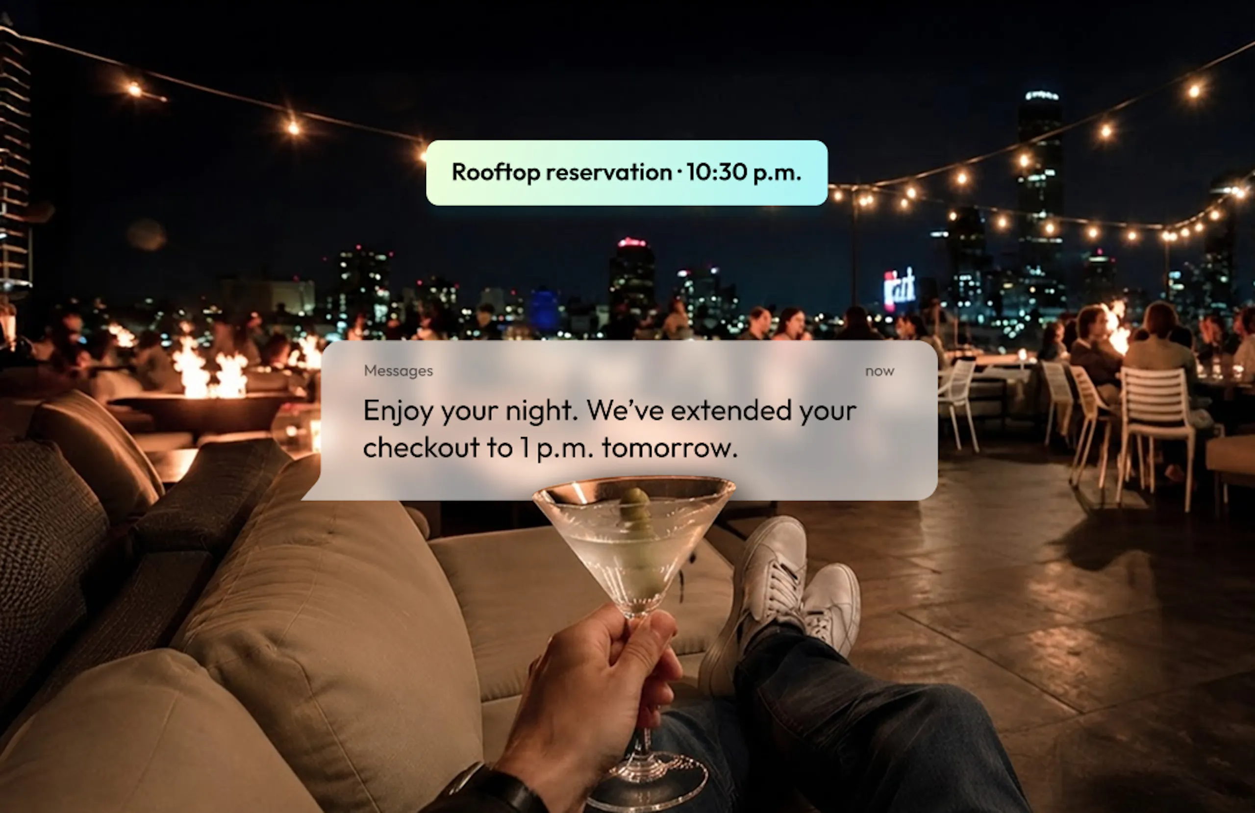 First-person POV of a person enjoying a martini on a rooftop bar; notification displays "Enjoy your night. We've extended your checkout to 1pm tomorrow."